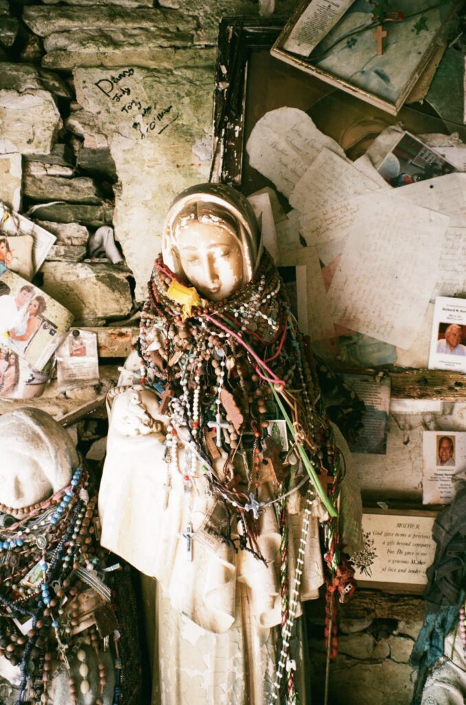 In front of a wall covered with photographs and written letters, a white sculpture in the shape of a woman wearing a long headscarf and robes has several beaded necklaces hung around its neck. A similar but shorter statue stands to its right.
