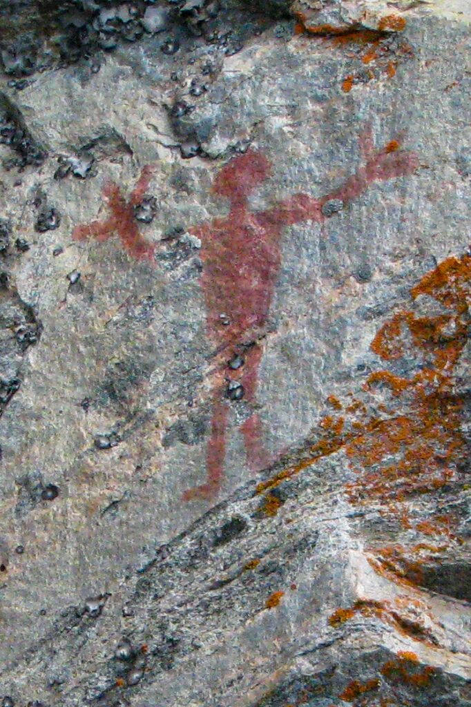 A close-up shows a red ochre rock painting on a rough stone surface. The painting depicts a human-like figure with arms outstretched.
