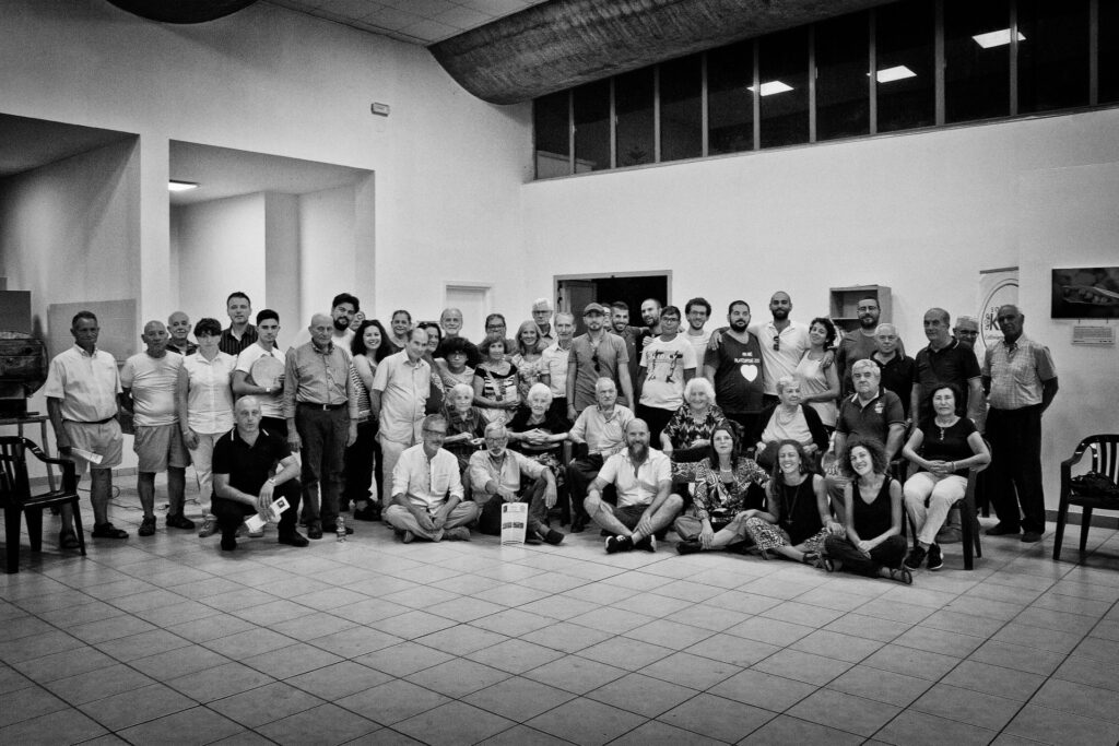 A black-and-white photograph shows a large group of people sitting and standing in rows in the corner of a room with high ceilings and a tiled floor.