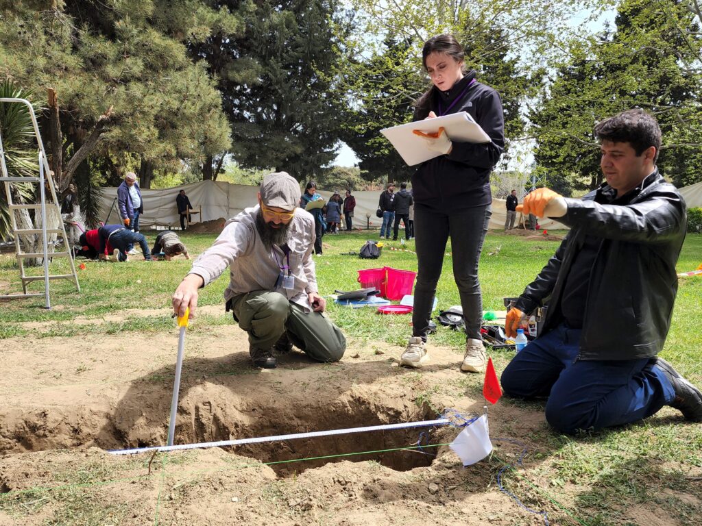 A person dressed in a black leather jacket and dark blue pants kneels beside a hole in the ground while a second kneeling person probes its contents with a long, slender metal tool. A third person clad in black stands and writes notes in a white binder.