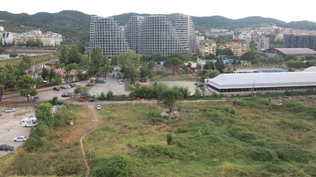 A small number of indistinguishable items surrounds a tree in a field in front of a long white structure, a large parking lot, a gigantic steel complex, and various pastel-colored buildings in a city.