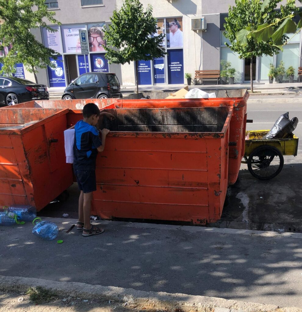 A child peers into one of two orange dumpsters in front of a motorbike with a yellow crate parked on a city street.