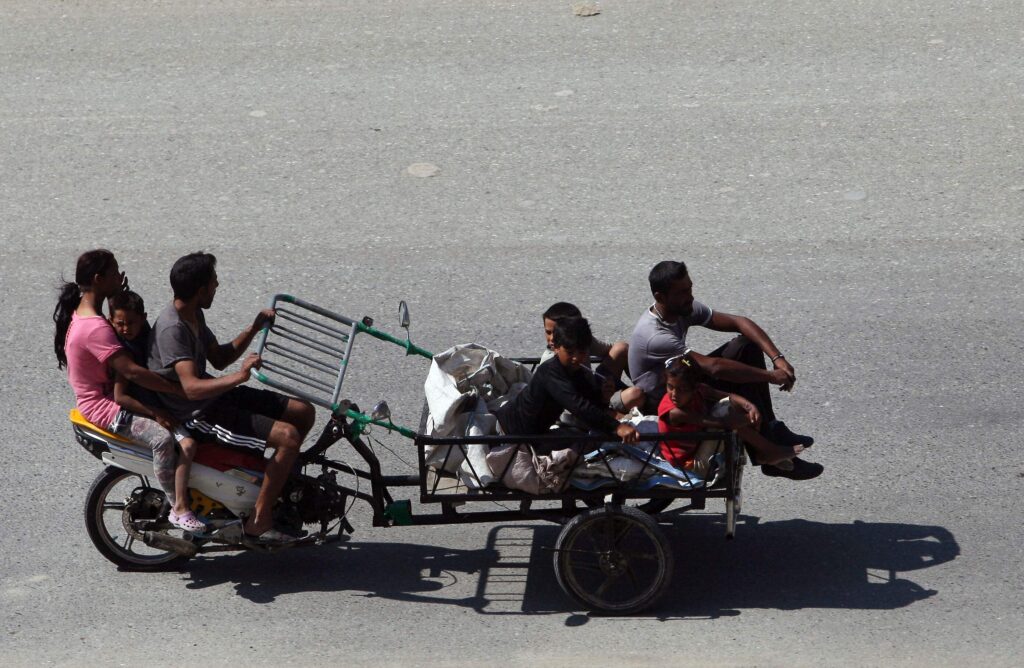 A woman, a child, and a man ride the back part of a motorbike, while the man holds a metal trailer attachment in front carrying four young people and large white sacks.