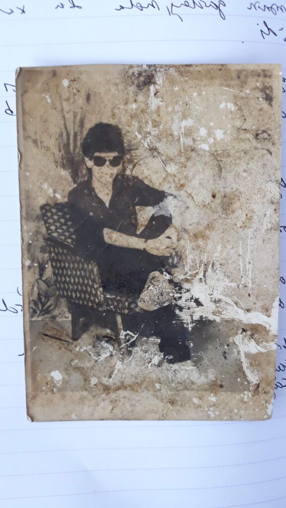 A weathered, blotchy black-and-white photograph depicts a person wearing sunglasses who sits in a chair with his legs crossed.