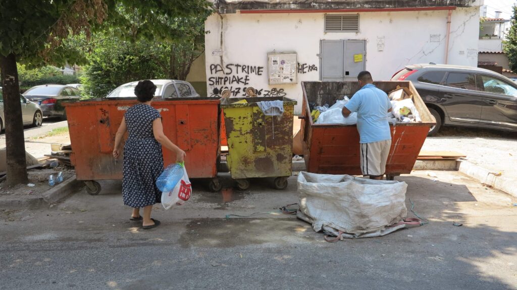 A woman wearing a navy-blue dress and carrying a blue plastic water bottle and a white plastic sack walks up to a man looking through one of three dumpsters next to large white sack.