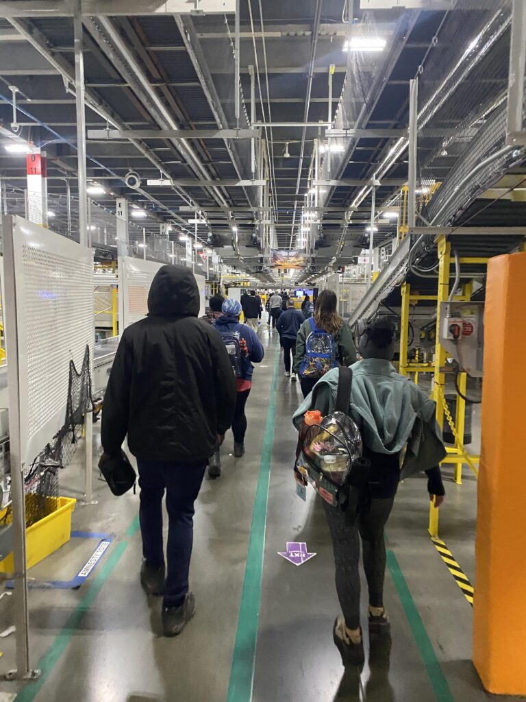 Two rows of people dressed in street clothes carrying backpacks walk down the narrow, brightly lit hall of a warehouse. The colors orange, yellow, green, and gray are prominent.