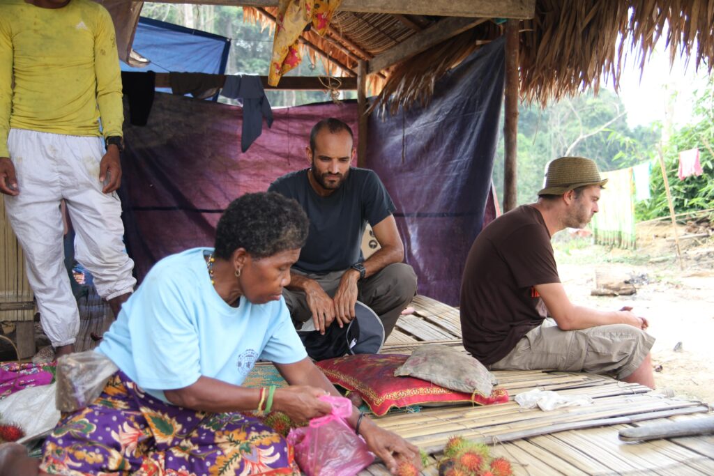 In an open shelter with bamboo floors and purple cloth walls, a man in a navy-blue T-shirt and gray pants squats and observes a woman in a light-blue T-shirt and purple, patterned skirt as she reaches for fruit. Two other people are also inside the shelter.