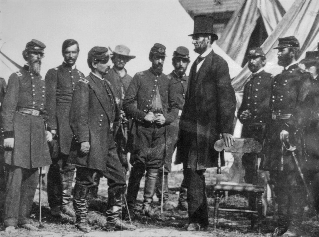 A black-and-white photograph shows Lincoln in profile as he visits with Union officers. He wears a black suit and his signature stovepipe hat.