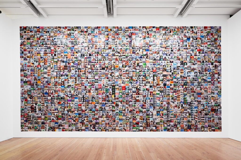 A museum installation shows a gallery wall composed of hundreds of photographs arranged side by side to form a colorful mosaic.
