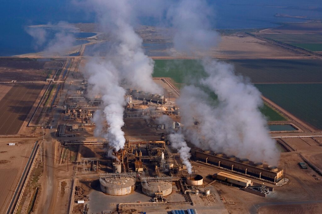 An aerial photo shows a huge industrial site spewing several plumes of steam, surrounded by green and brown fields, with a blue sea in the background.