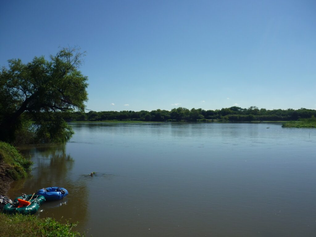 Two small inflatable boats float by a tree next to the bank of a river with a calm, lightly rippled surface. Trees are visible on the opposite bank and a cloudless blue sky stretches overhead.