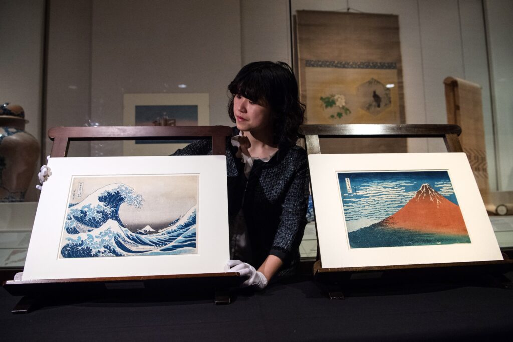 A person dressed in black wearing white gloves holds up a matted picture of a cresting blue wave. Beside them is another matted picture of a red volcano.