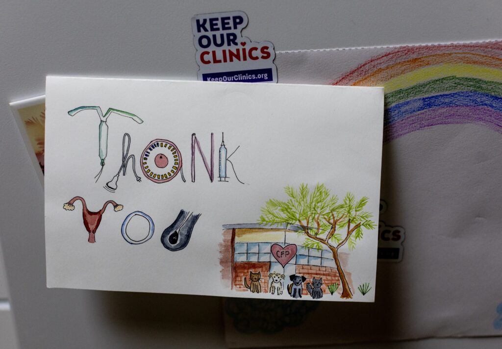 A hand-drawn “Thank You” card whose letters are formed out of reproductive health-themed images hangs on a surface near a magnet that says: “Keep Our Clinics.”