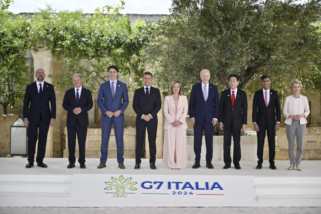 World leaders of varying heights stand next to one another in front of green foliage. A banner in front of them reads “G7 Italia 2024.”
