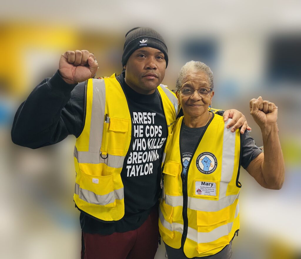 Two people wearing yellow reflective vests stand arm-in-arm holding their opposite fists aloft.