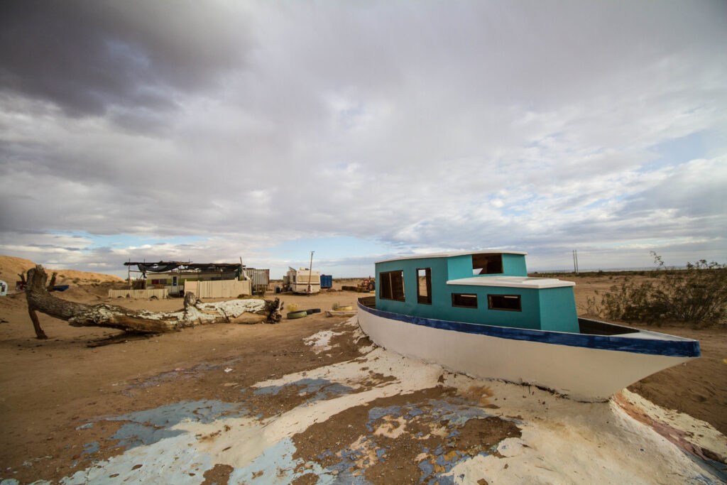 The shell of a turquoise, blue, and white boat and a derelict building stand in a dusty, barren landscape.