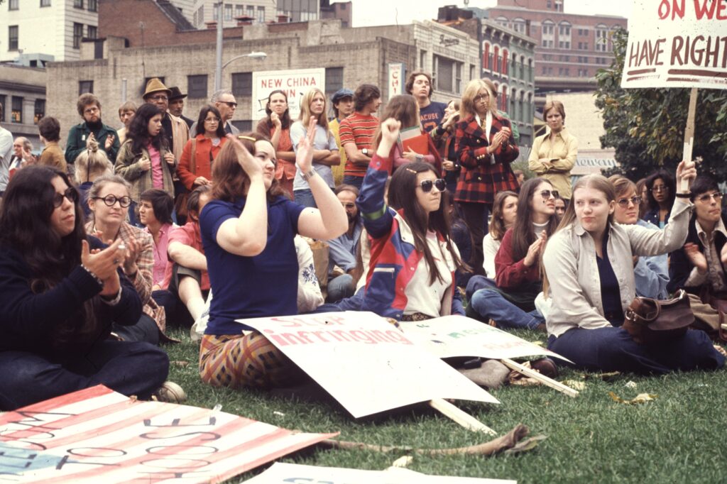A large group of women and some men sit on a green lawn holding signs and clapping their hands.