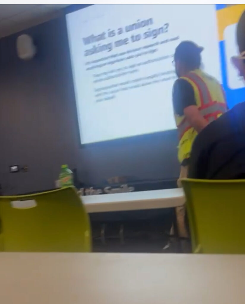 A person wearing a yellow and orange reflective vest stands in a room with yellow chairs. Text projected on a screen reads: “What is a union asking me to sign?”