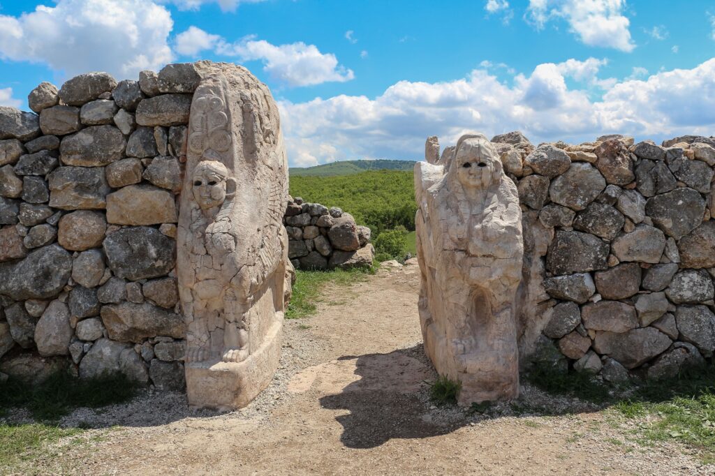 A brown path runs between stone-carved figures that form the entrance of a gate. Beyond the gate is green grass and blue sky with white, fluffy clouds.