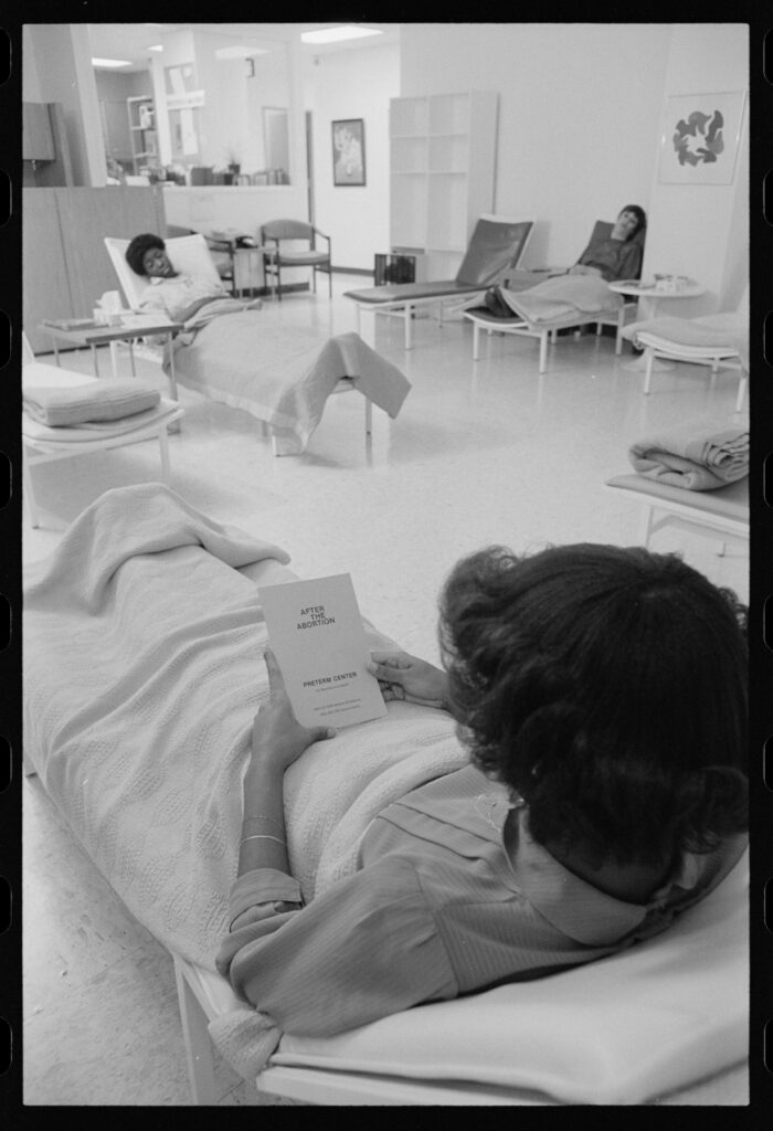 A black-and-white photograph shows three people in a clean white room sitting on reclining chairs. One person holds a pamphlet titled “After the Abortion.”