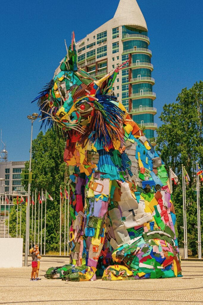 Two people look up at a large, multicolored statue of an Iberian lynx. Behind the statue is a high-rise building, trees, and cloudless blue sky.