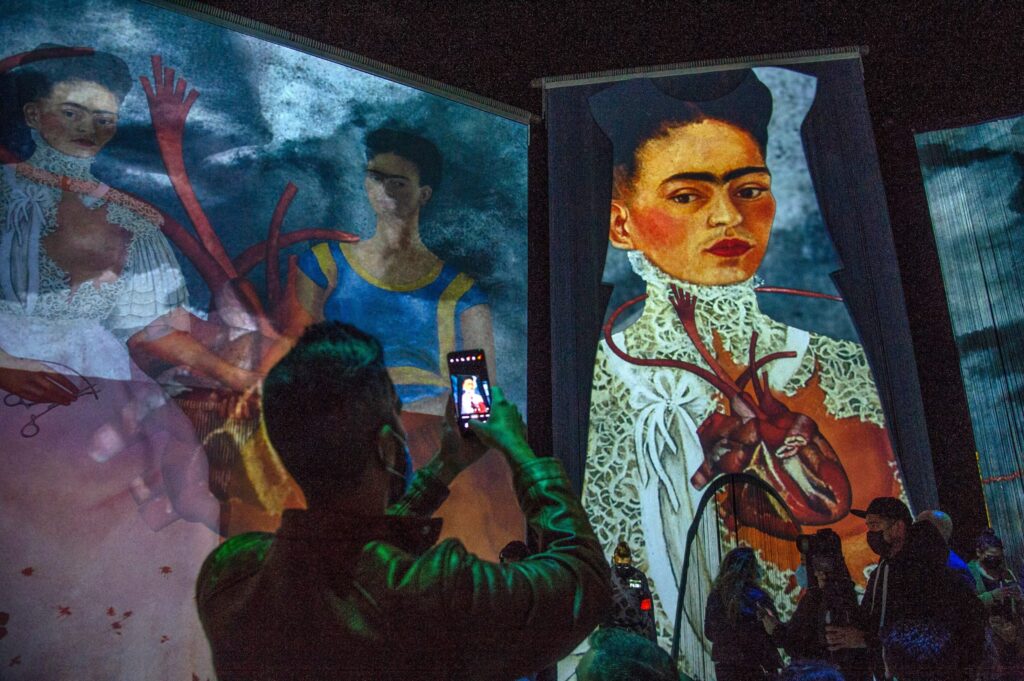 A person holds up their smartphone to take a picture of a large colorful portrait projected on a wall. Blue, purple, green, white, black, and orange hues predominate.