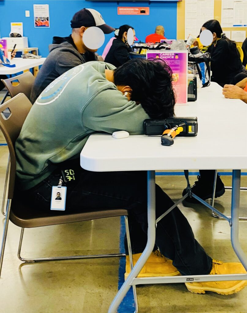 A person wearing a teal sweatshirt and earbuds has their head in their arms on top of a table.