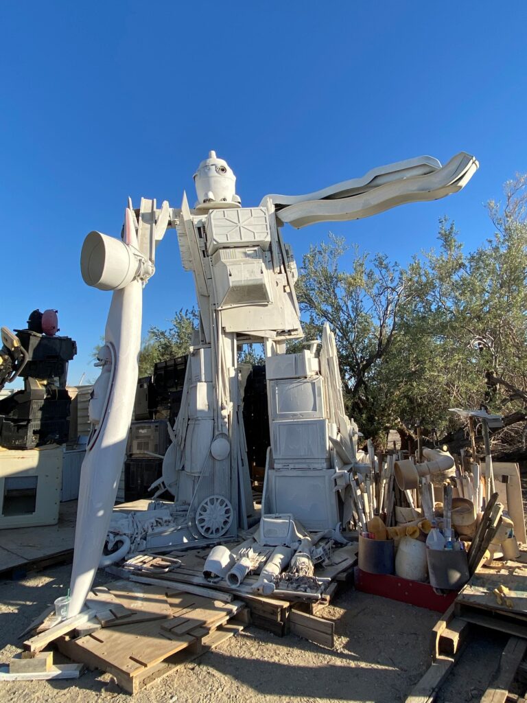 A large robot statue made from discarded appliances and other objects painted white stands next to old tools, wooden pallets, and indistinguishable waste.