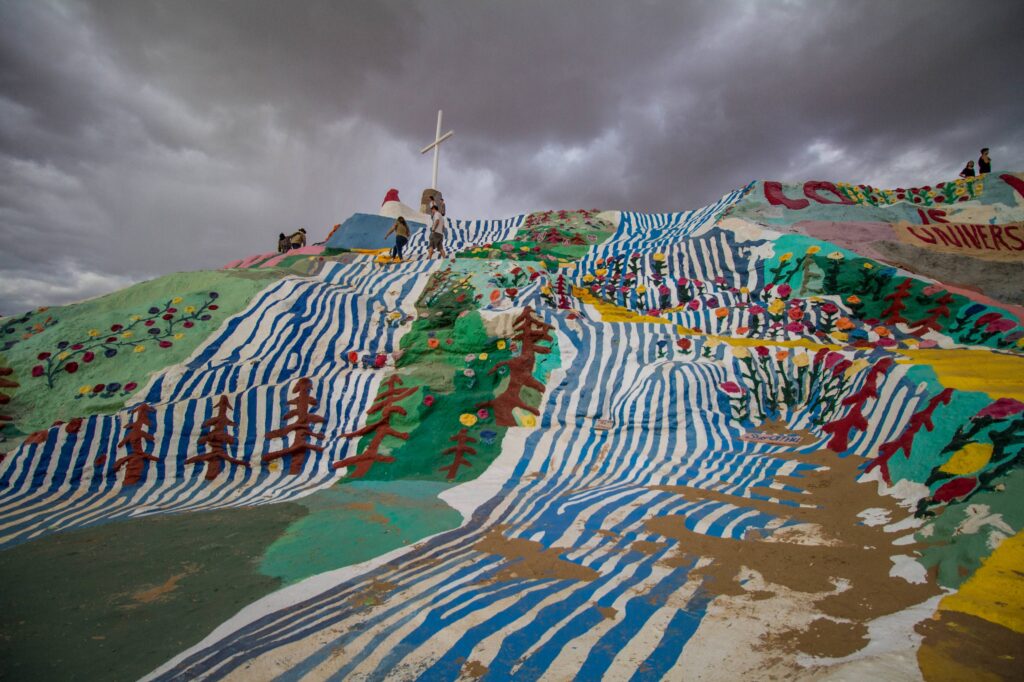 Four people walk atop a huge hill-like construction painted with blue stripes resembling rivers, brown trees, and multicolored flowers.