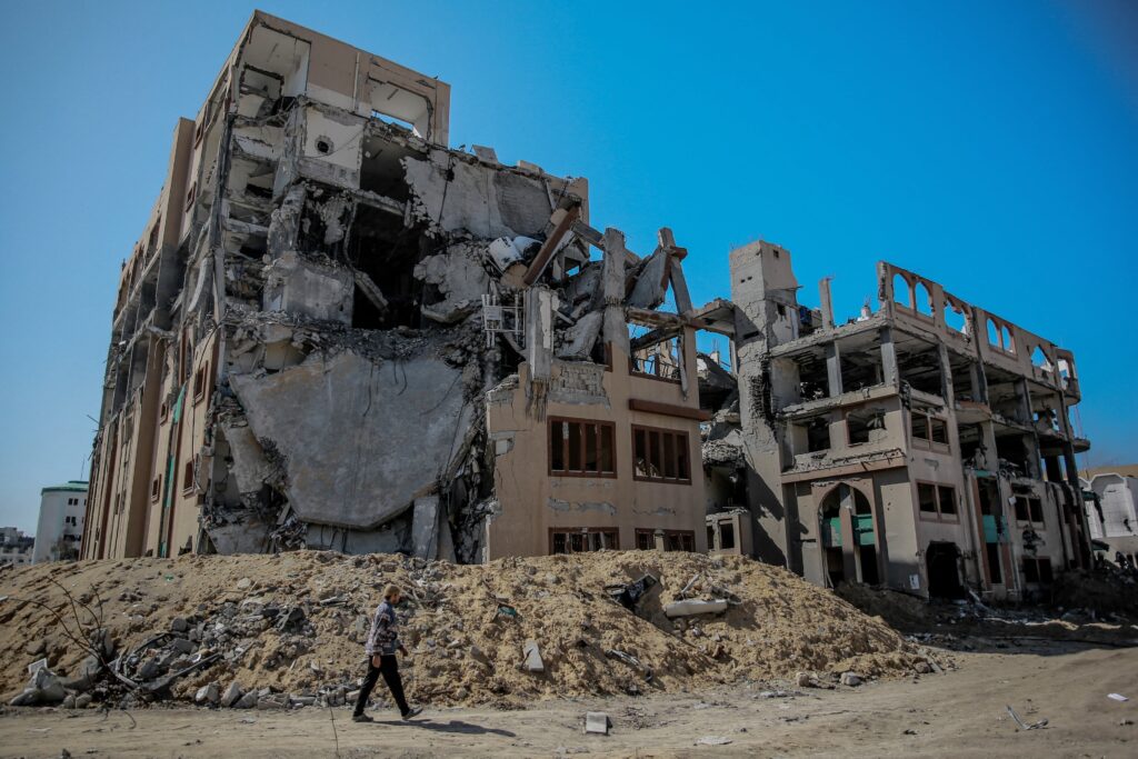 Under a cloudless azure sky, a person walks in front of a bombed-out building. A pile of rubble surrounds the building’s base.