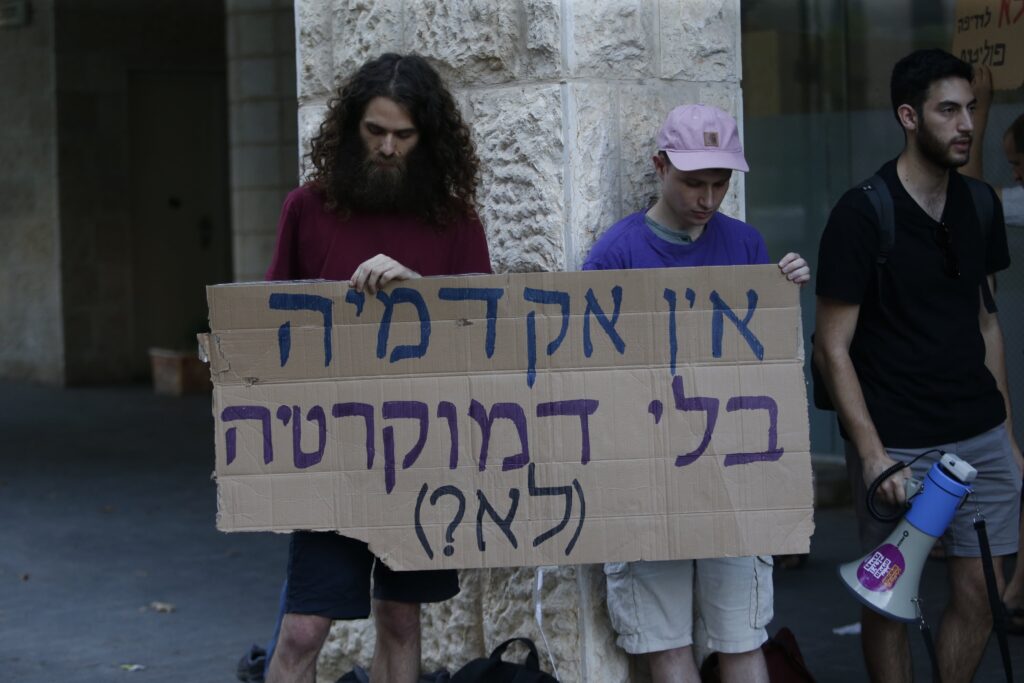 Two demonstrators lean against a pillar while holding a sign with Hebrew writing. A third stands to their left holding a bullhorn.