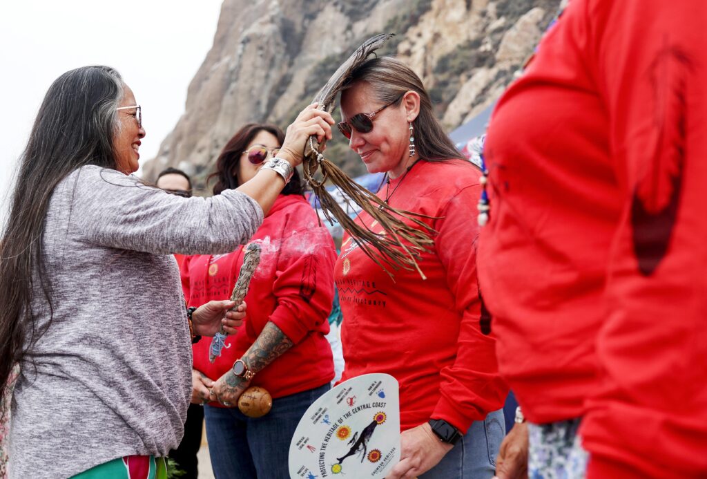 A group of people stand on a beach. One person with long dark hair and a gray shirt touches a second person, dressed in red, on the head with a traditional object made of feathers and leather tassels.