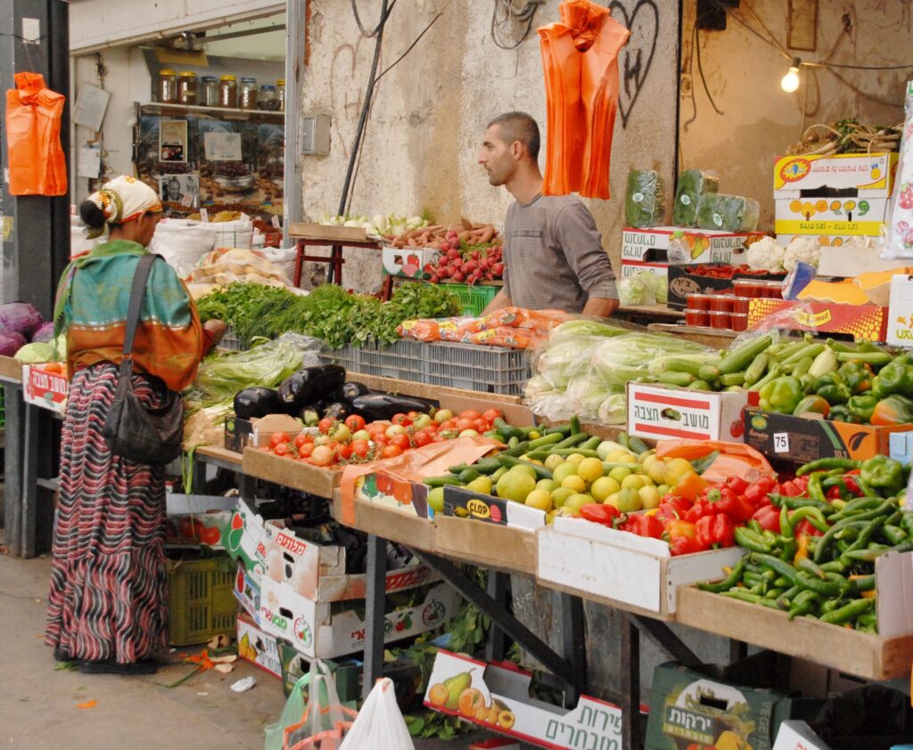 A person wearing a skirt and headscarf shops at a market stall attended by a person standing behind bins of brightly colored vegetables for sale. Red, green, orange, yellow, and beige colors are prominent.