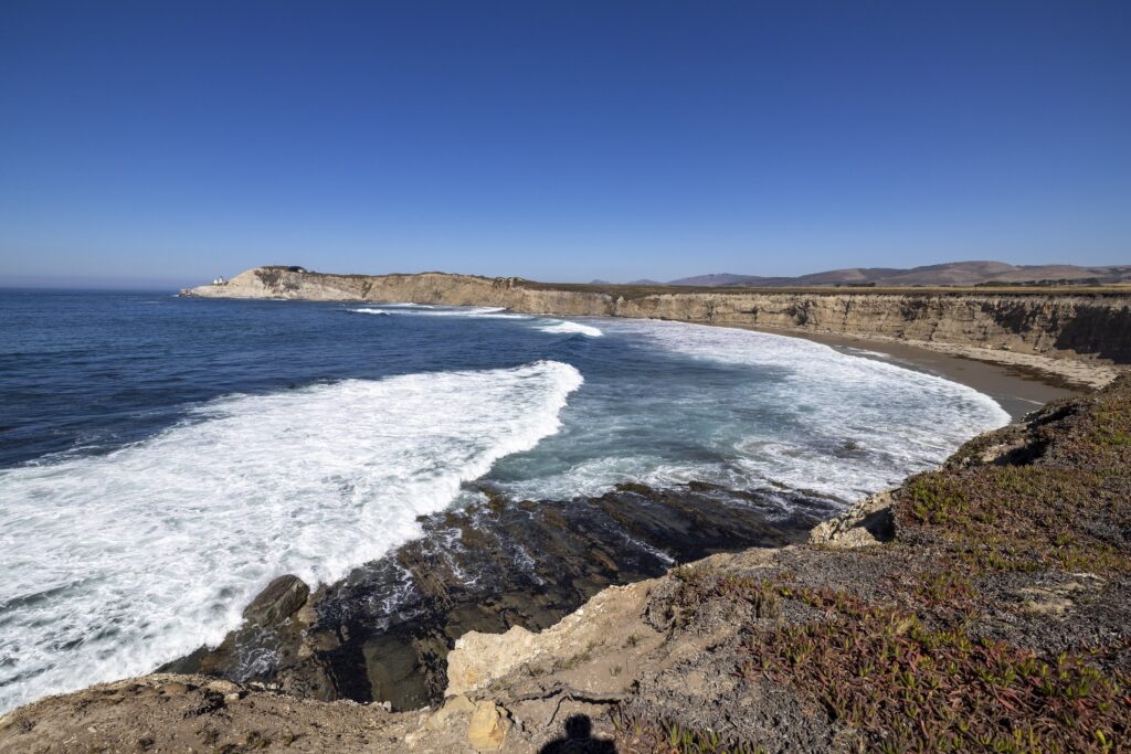 Foamy white waves wash against a tan-colored coastal area. They sky overhead is a deep blue that lightens in color as it meets the horizon.
