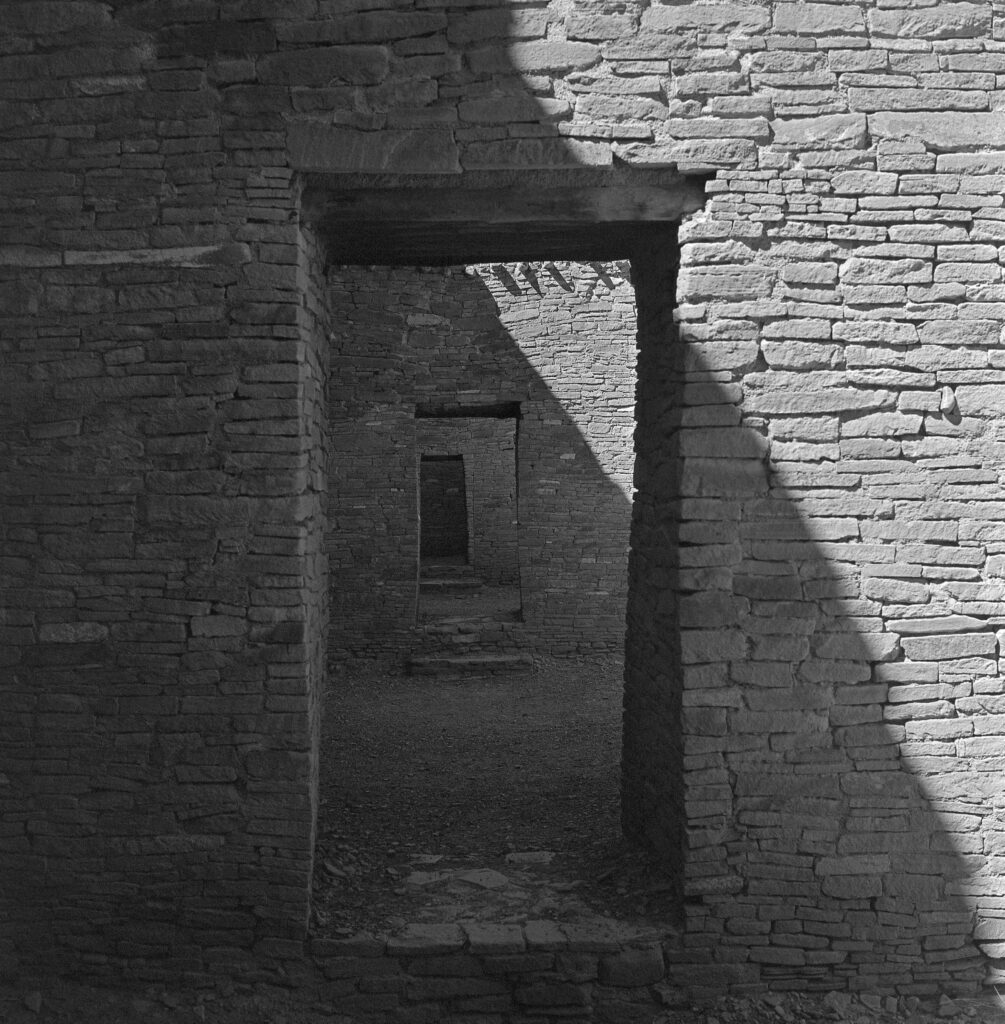 A black-and-white photograph shows multiple entryways in carefully built sandstone walls that lead into one another. Shadows cast on the walls create an interplay of dark and light.