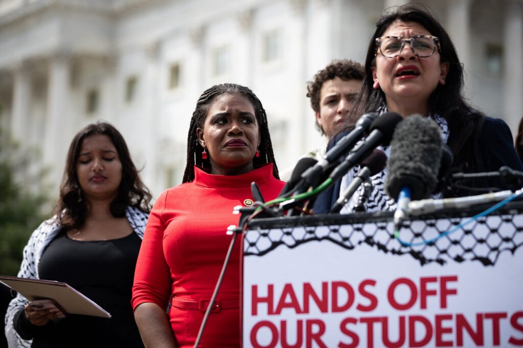 A woman wearing glasses speaks into microphones at a podium with a sign that says: “Hands off our students.” Three other people stand to her right.