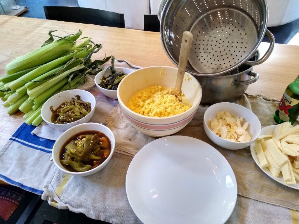 Ingredients and equipment for cooking—including corn, butter, and peppers, plates, bowls, and a pot—are laid out on a countertop.