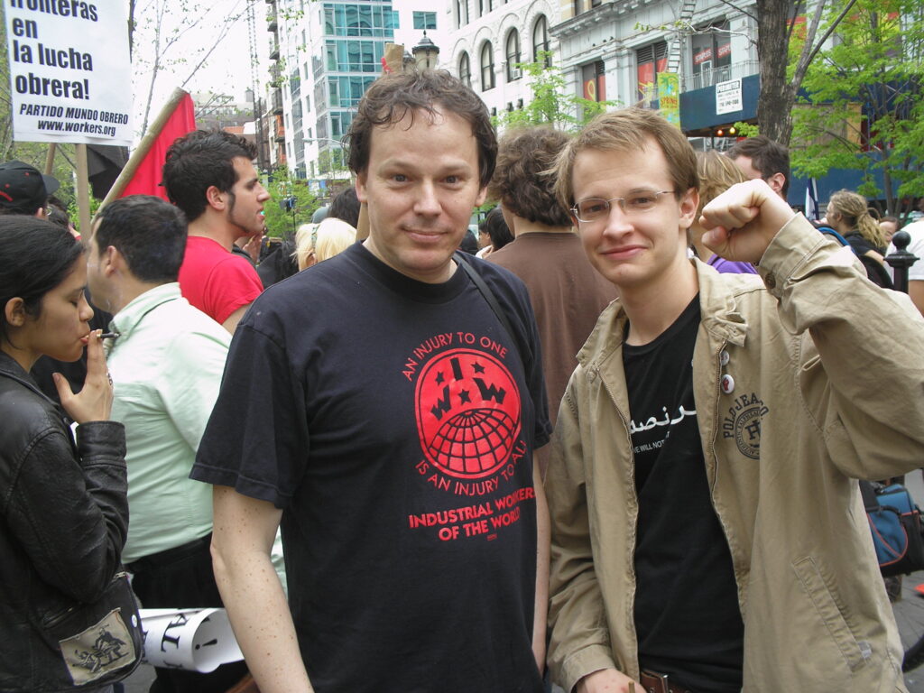 On a street filled with a group of people, two people pose for the camera, with one raising his fist. They both wear black T-shirts.