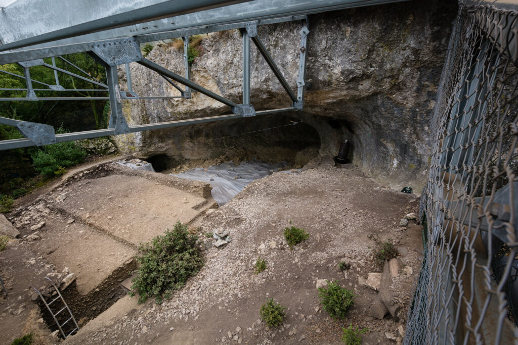 A fence on the right and a metal structure above frame the opening of a cave under excavation.