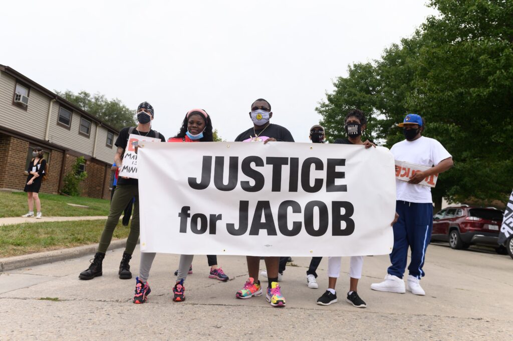 A group of people stands on the pavement near residential housing holding a white sign with black letters that reads “Justice for Jacob.”