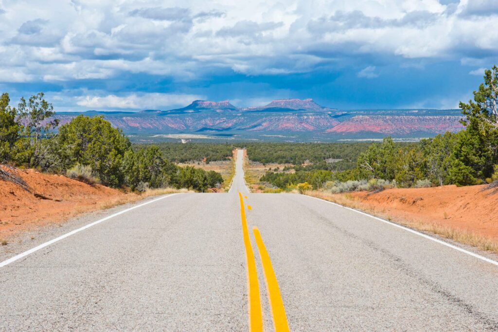 Under a cloudy sky, a road bordered by red dirt and low shrubs with a yellow line down the center stretches far into the distance, where two distinctive sandstone formations are visible.