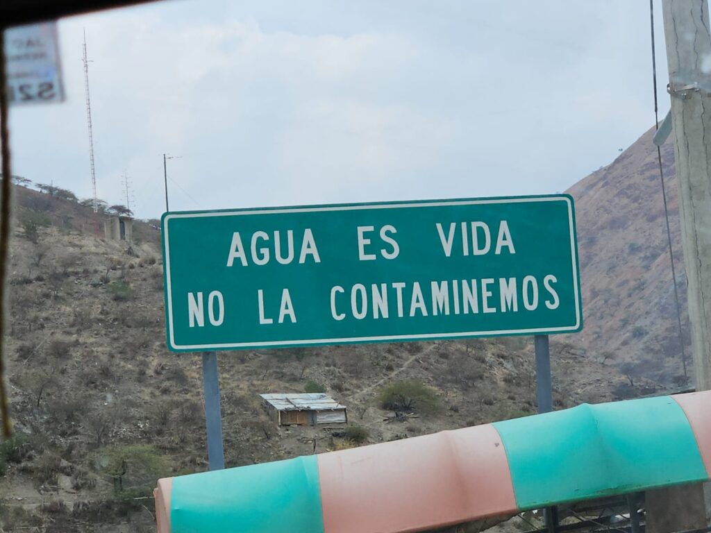 A green road sign with white letters and a white border stands amid greenish-brown hills. A green and pink tube stretches in front of it.