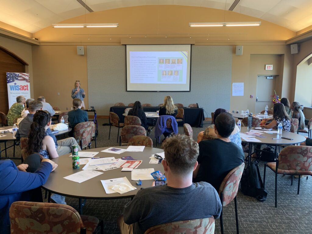 People sit at round tables looking toward a screen next to which a presenter with a microphone speaks.