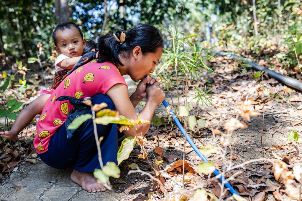 A woman with a small child on her back crouches among tan dirt and green shrubs, holding onto a thin blue hose.