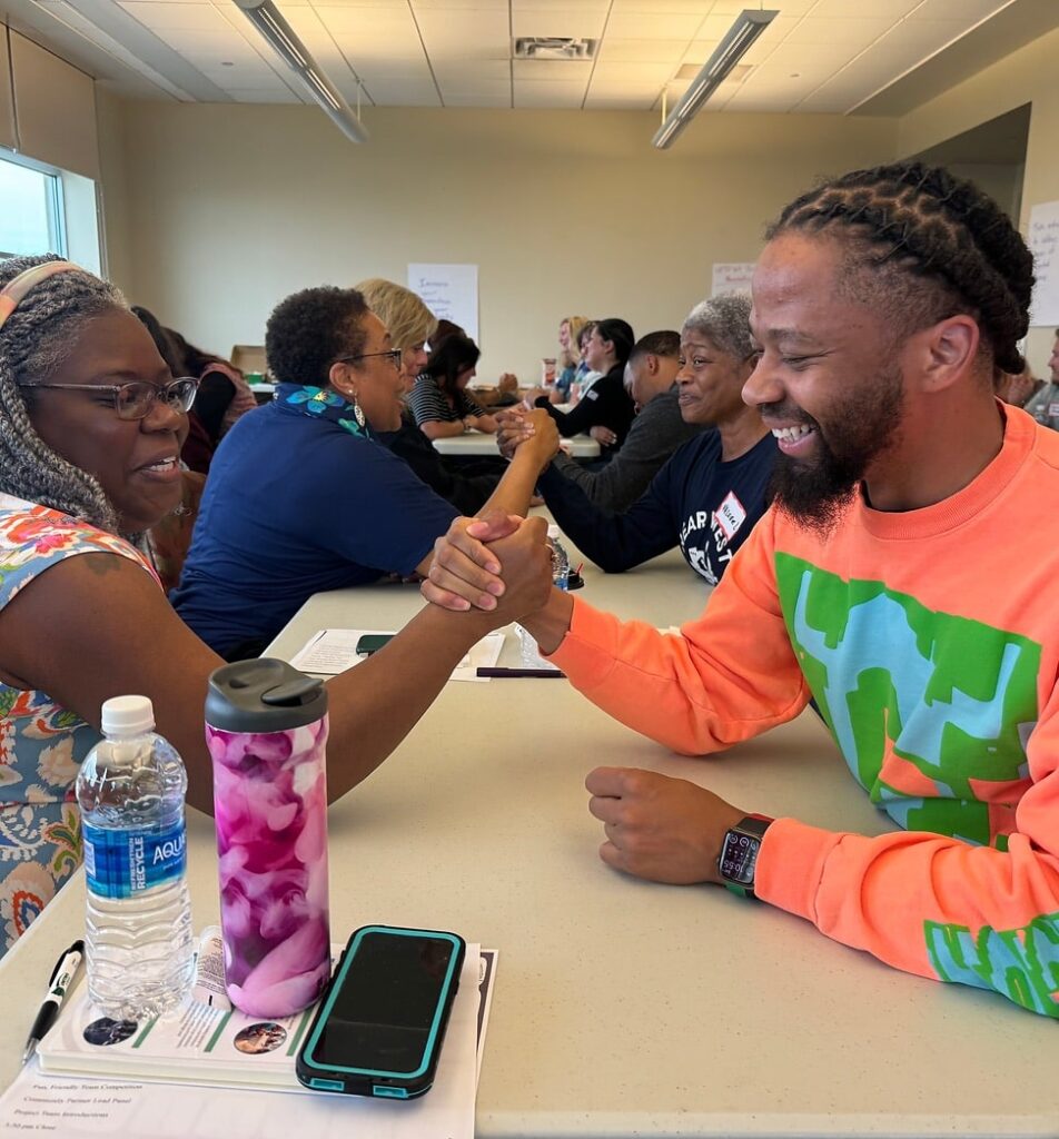 People at a long table sit across from one another and clasp hands in the style of an arm-wrestling match.