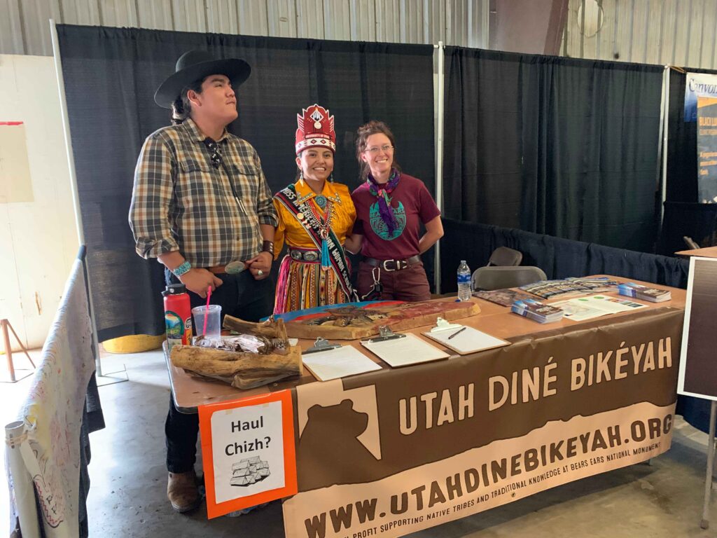 Three people wearing a plaid shirt and jeans, a decorative yellow shirt and multicolored skirt, and a T-shirt and dark-red jeans stand behind a table.