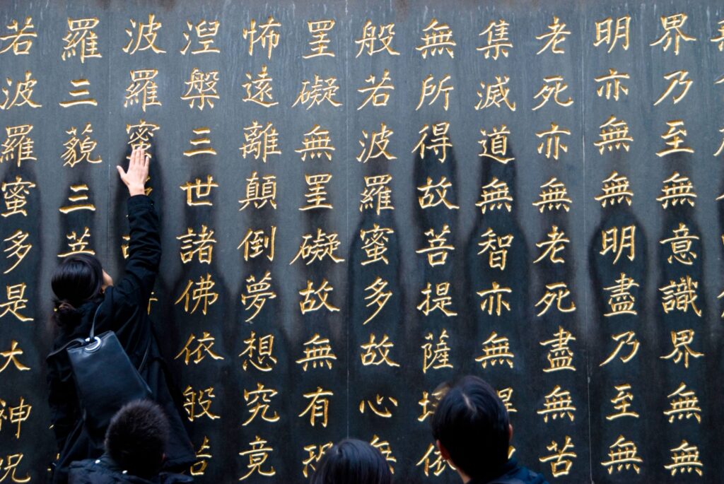 Em detalhe de foto mostrando uma parede de pedra gravada com grandes caracteres dourados, uma pessoa se aproxima para tocar a escrita enquanto outras observam.