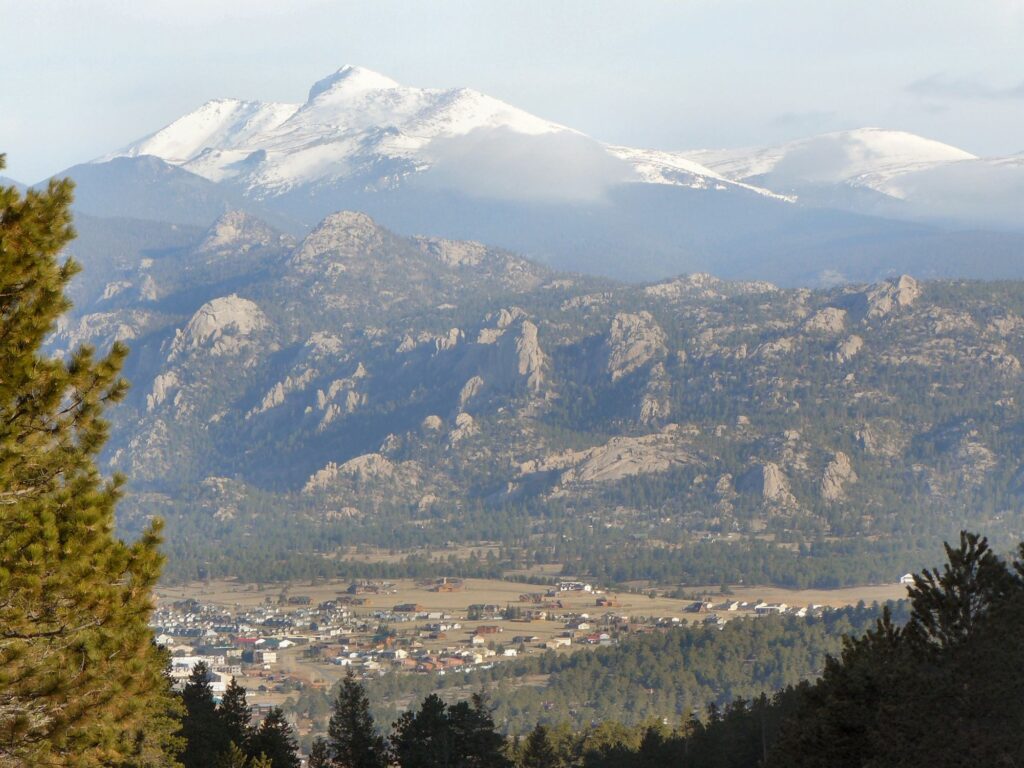 A wide view shows a mountainous landscape, featuring snow-capped peaks in the background and rugged rock formations in the foreground. Residential houses and other buildings nestle in the valley below.