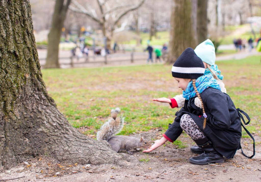 Two children dressed in knit hats, scarves, and jackets crouch near the base of a tree, reaching out their hands toward a small brown animal with a fuzzy tail.