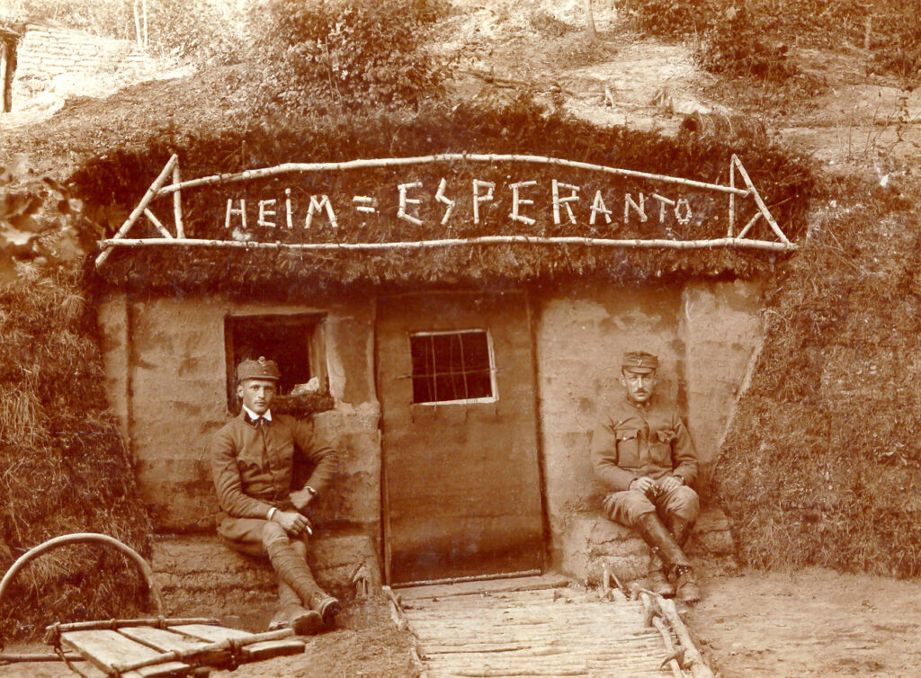 Duas pessoas de uniforme sentadas do lado de fora de um abrigo com as palavras \“HEIM = ESPERANTO\”gravadas acima da porta.